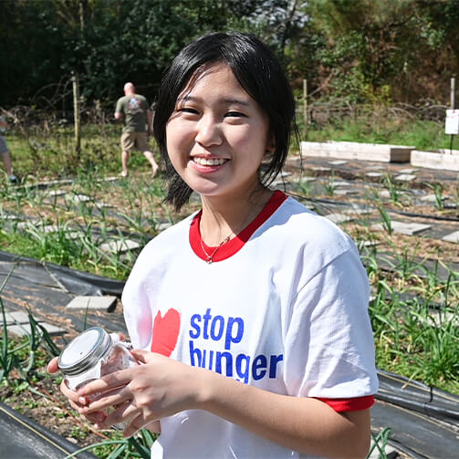 A young woman holds a jar of food while standing in a vibrant garden filled with plants and flowers.