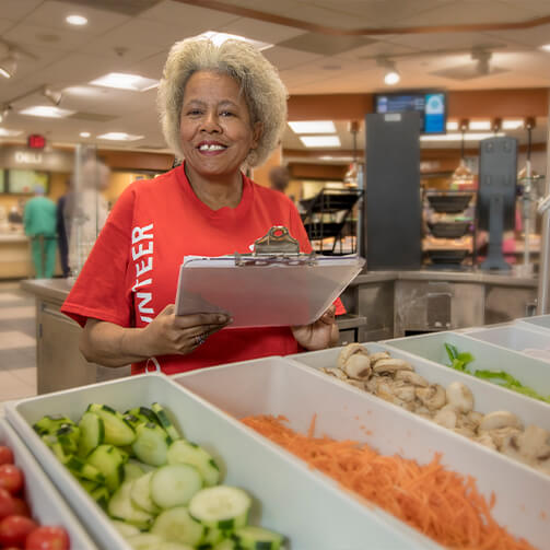 Smiling woman wearing a red “Volunteer” t-shirt holding a clipboard in a cafeteria, standing in front of a salad bar with various fresh vegetables.
