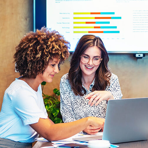 Two women collaborating at a laptop with a data chart on a screen behind them