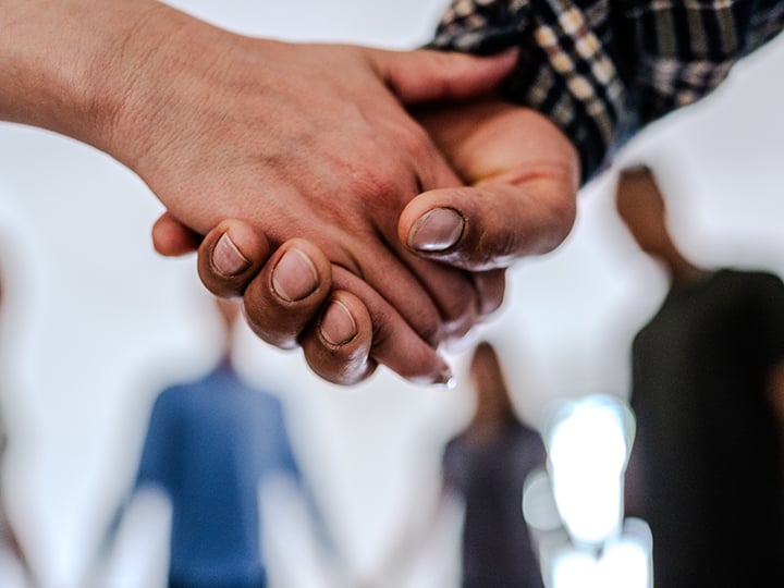 A close-up of two hands clasped in support, with a blurred group of people standing in a circle in the background.