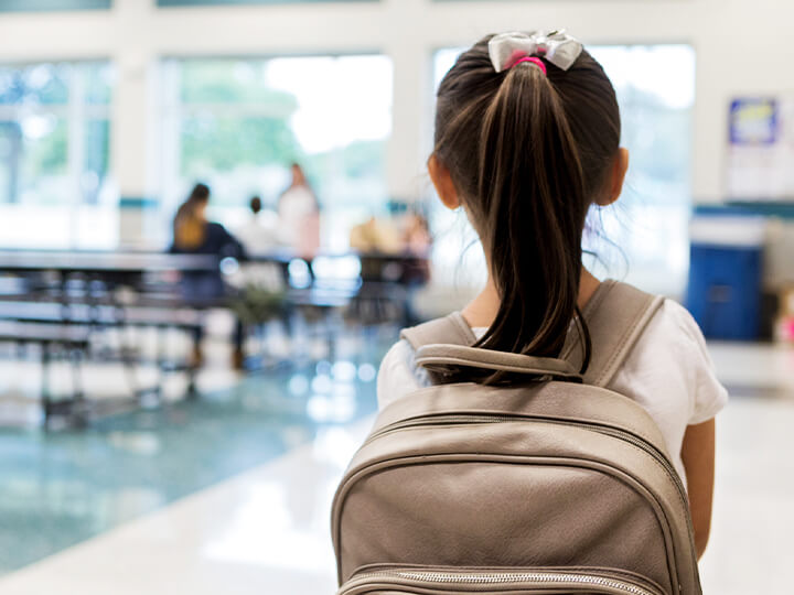 Image of a school cafeteria with a young person standing in the foreground. The individual, wearing a ponytail and a backpack, looks towards a group of people sitting at tables in the well-lit background. Large windows fill the space with natural light, creating a bright and welcoming atmosphere