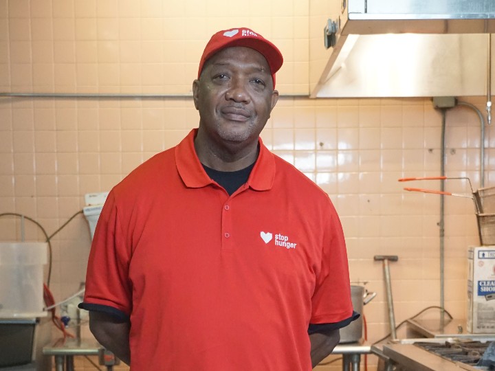 Person wearing a red polo shirt and red cap with a white heart logo and 'stop hunger' text, standing in a kitchen with tiled walls and various kitchen equipment in the background