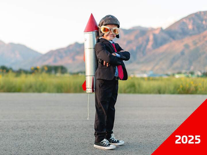 Child wearing a suit, tie, and helmet stands on an open road with a homemade rocket strapped to the back, mountains in the background, and 2025 in bold white text on a red corner banner.