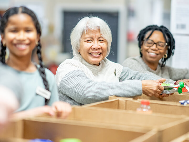 Three individuals collaboratively packing cardboard boxes with various items, including a green packaged product and a red bottle, possibly in a volunteer or community service setting
