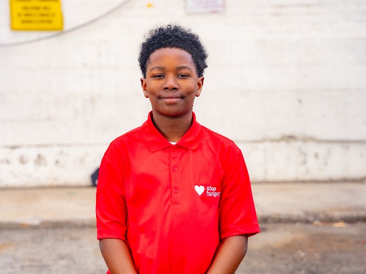 Person wearing a red polo shirt with a white heart logo and 'stop hunger' text, standing outdoors near a concrete wall with a yellow sign in the background, likely part of a hunger relief campaign