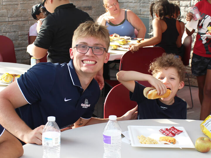 A man and a boy enjoying hot dogs together at a picnic table, smiling and sharing a moment of joy.