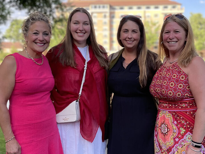 Four women smiling and posing outdoors in colorful dresses, standing close together with trees and a building in the background.