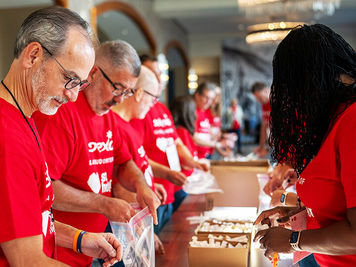 Volunteers in red shirts collaborate at a table, preparing care packages in a large indoor setting with chandeliers and arched doorways, highlighting teamwork and community service.