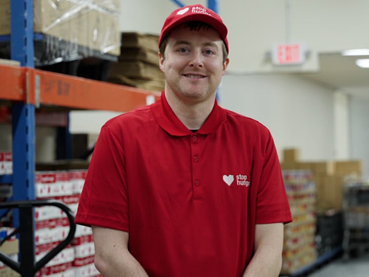 Portrait of a person wearing a red Stop Hunger polo shirt and matching cap, standing in a warehouse aisle with shelves, pallets, and stacked food boxes in the background.