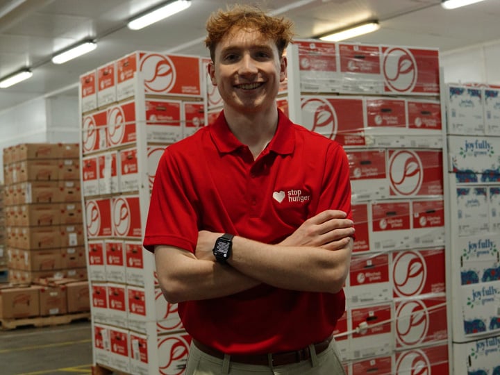 Portrait of a person wearing a red Stop Hunger polo shirt, standing with arms crossed in a warehouse aisle with stacked food boxes on pallets and bright overhead lighting.