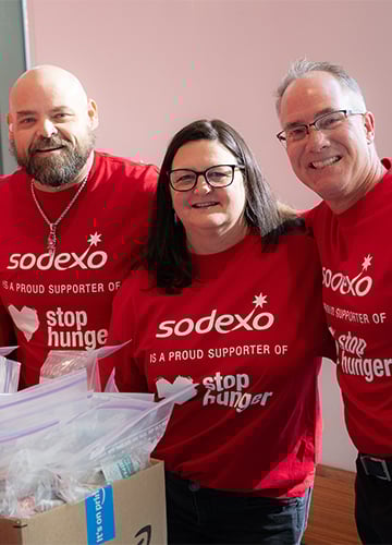 Three smiling people in red Sodexo "Stop Hunger" shirts stand together near a box of care packages.