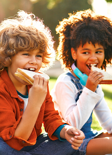 Two children sitting on green grass, happily eating sandwiches during a sunny day.
