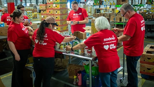 Group of people in red Sodexo Stop Hunger shirts work together in a warehouse or food bank, organizing and packing canned goods on a metal table amid boxes and shelves.