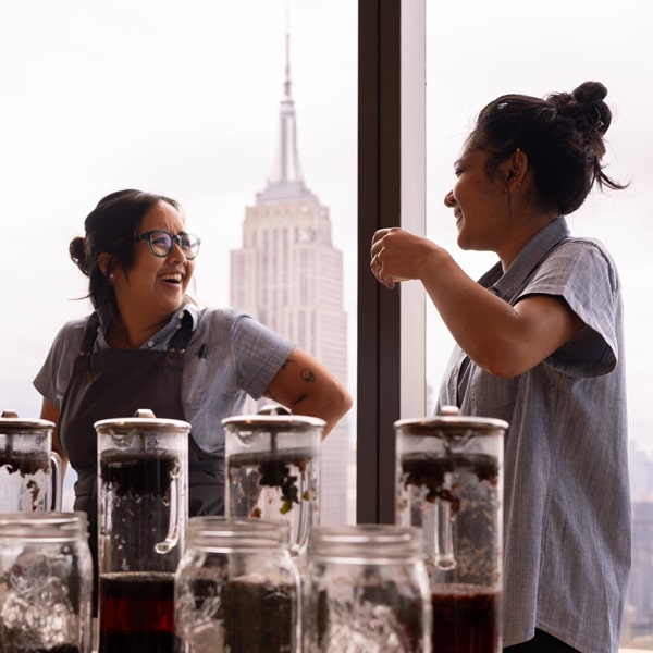 Two women stand by a window, gazing at the Empire State Building in the background, showcasing a vibrant city view