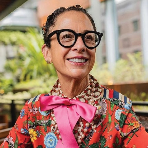 Person in glasses and floral outfit with pink bow, seated outdoors with greenery in the background.