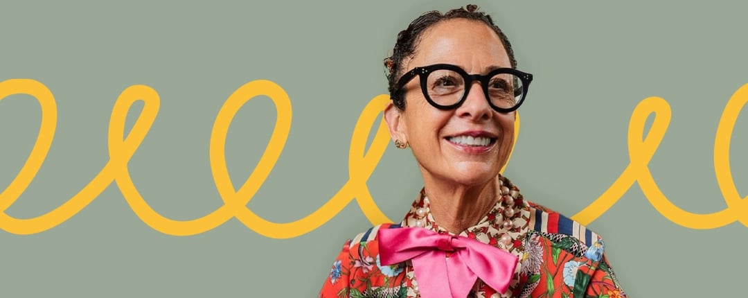 Person wearing large black glasses, a colorful floral shirt with a pink bow tie and patterned collar, set against a light green background with a yellow squiggly line across it.