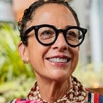 Person with curly hair and glasses smiling outdoors in a patterned shirt.