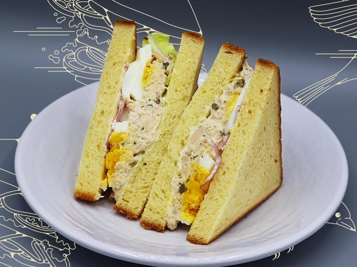 Plate with two upright triangular sandwich halves made with toasted bread, filled with tuna salad, hard-boiled egg slices, lettuce, and herbs; background features abstract decorative patterns.