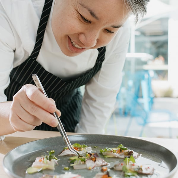 Person in white shirt and black apron using tweezers to plate gourmet food in a bright kitchen with blue chairs and tables.