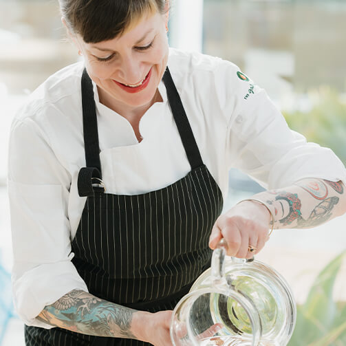 A woman in a white chef's coat and black pinstriped apron, showcasing tattooed arms while pouring liquid from a glass pitcher in a professional culinary setting.