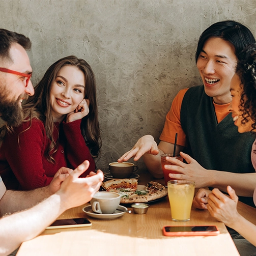 Four people sit around a wooden table sharing a meal with pizza and drinks including coffee and juice; background features a plain wall.