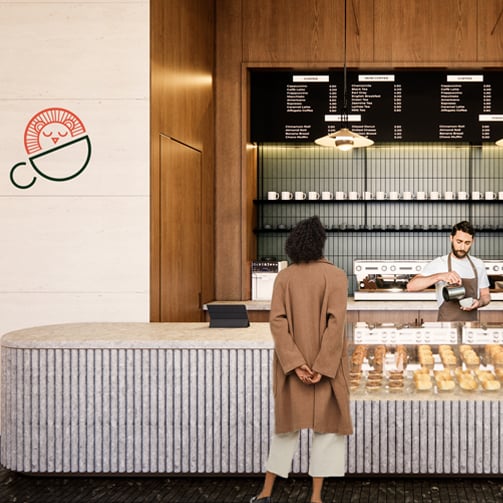 Customer standing at a modern café counter while a barista prepares a drink behind a glass pastry display, with menu boards and a minimalist logo on a wood-paneled interior wall.