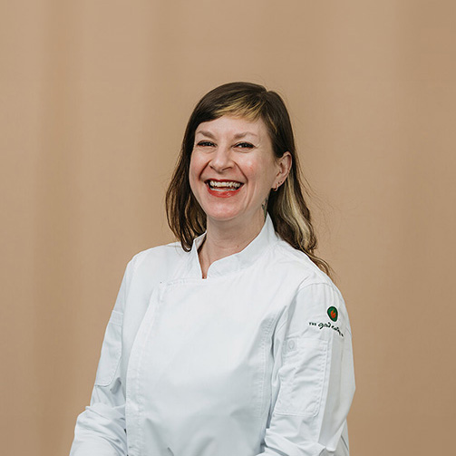 Professional female chef in a white jacket with a logo on the sleeve stands against a plain beige background.