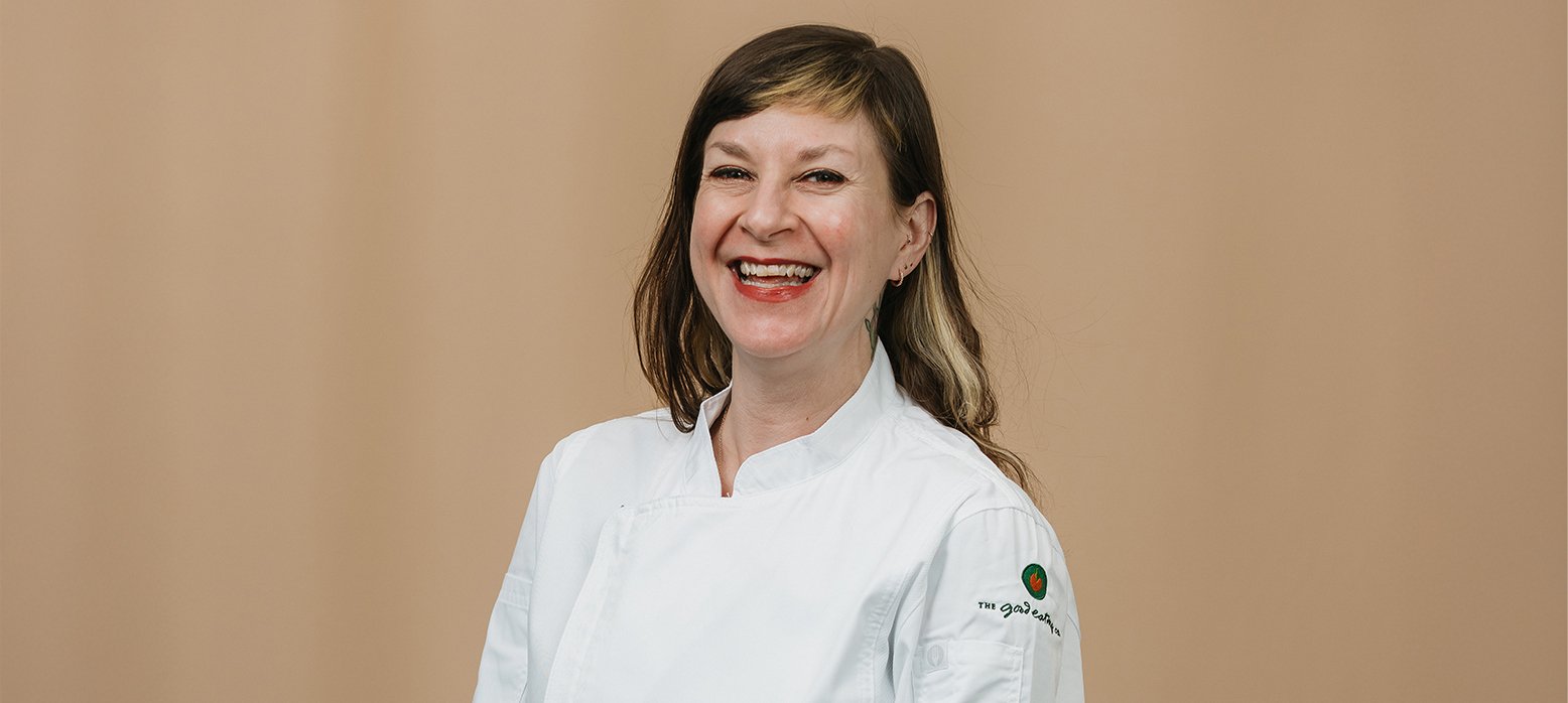 A woman wearing a white chef's coat with a blurred face, featuring the logo 'THE GOOD EATING' on the left sleeve against a plain beige background.