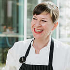 A professional female chef wearing a white chef's coat and black apron stands in a kitchen setting, with glass windows and kitchen equipment visible in the background.