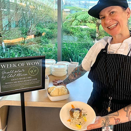 A chef stands next to a table holding a plate of food, with a sign promoting a 'Week of Wow' event featuring small plates by Chef Alicia, including a vegan Lion's Mane Cake with Chickpea Remoulade