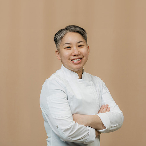 Female in a white chef's jacket stands with arms crossed against a beige background.