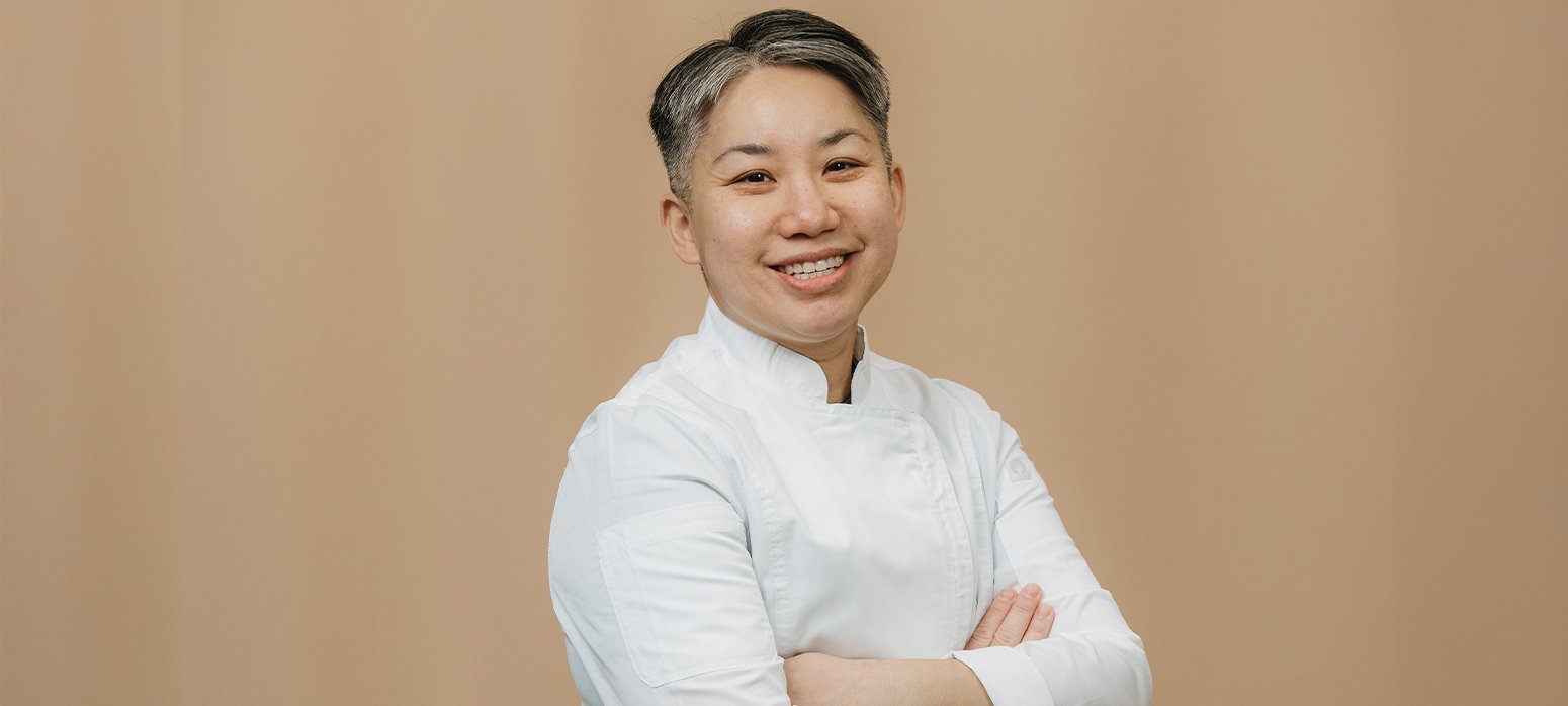 A woman dressed in a white chef's jacket with arms confidently crossed, set against a plain beige background. Their face is intentionally blurred for anonymity.