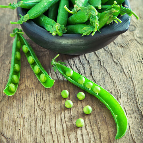 A bowl filled with vibrant green peas, accompanied by two open pea pods and scattered loose peas on a wooden surface.