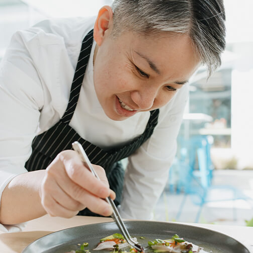A female chef in a white shirt and a black apron with white stripes, carefully plating food using tweezers in a bright, modern dining setting