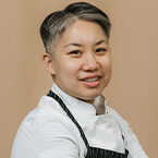 A woman wearing a white shirt and a black apron with vertical stripes, standing against a plain beige background, possibly portraying a culinary professional