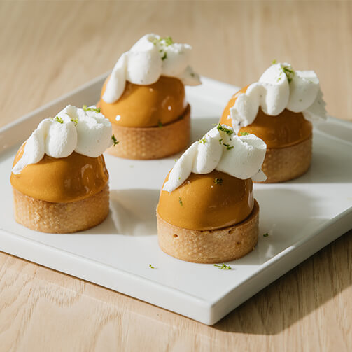 Four small pastries on a white square plate, each featuring a caramel-colored dome filling topped with whipped cream and garnished with green zest, elegantly arranged as a dessert.