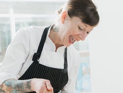 A woman with tattoos skillfully prepares food in a kitchen, showcasing her culinary talents and unique style.