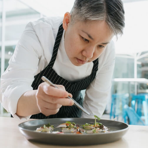 A woman in a white apron skillfully prepares food on a plate in a bright kitchen setting.