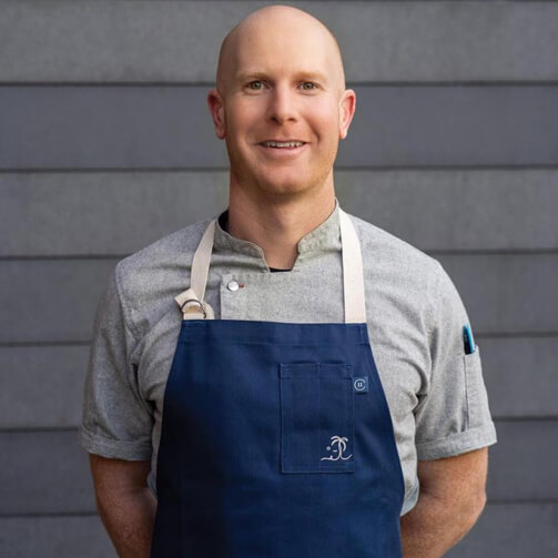 A bald man in an apron stands against a gray wall, ready for cooking or preparing food.