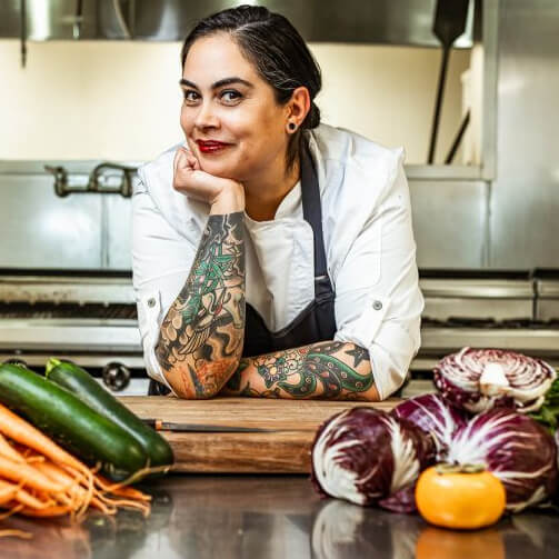A woman with tattoos sits at a table surrounded by an array of fresh vegetables.