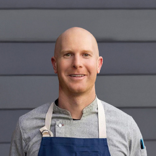 Male chef in a gray shirt and blue apron stands in front of a gray wall.