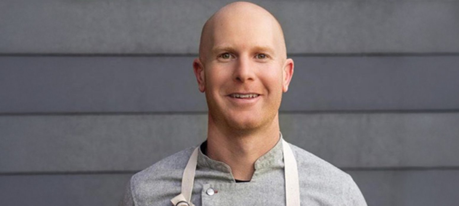 Male chef in a gray shirt and blue apron stands in front of a gray wall.