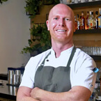 A chef in a grey apron and white coat stands confidently with arms crossed, set against a backdrop of shelves filled with bottles and glasses in a bar or restaurant.