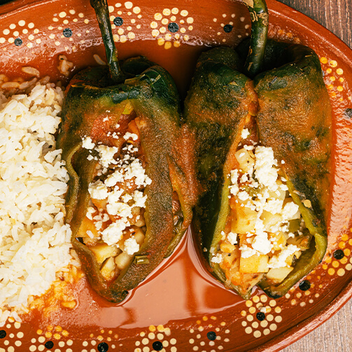 "Two stuffed poblano peppers topped with crumbled cheese, served alongside white rice on a colorful plate.