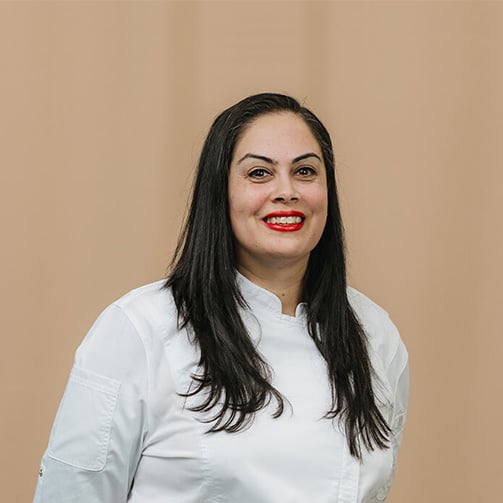 Woman wearing a white chef's jacket, standing against a plain, light brown background with their face blurred for anonymity