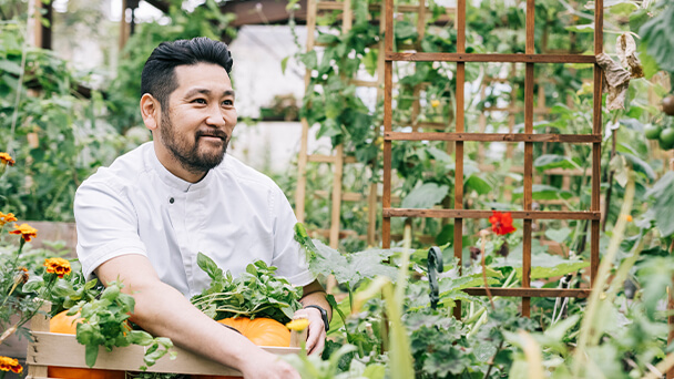 A man in a white shirt holds a basket filled with fresh vegetables, showcasing a vibrant assortment of produce.