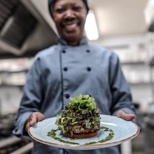 Chef in uniform holds a plated gourmet dish with stacked greens and a base, garnished with green sauce, in a professional kitchen setting.