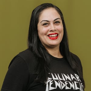 Person with long black hair wearing a black t-shirt  against a solid olive green background.