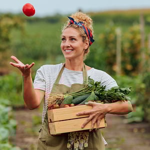 Person in a garden juggles a tomato while holding a crate of cucumbers, leafy greens, and other vegetables.