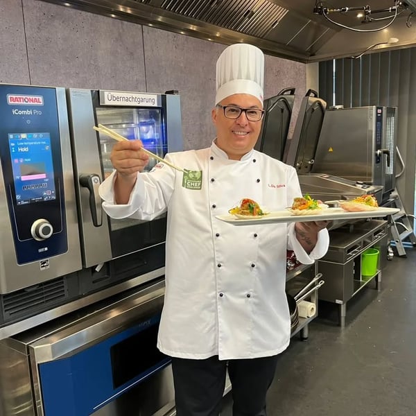 Chef sorridente segura prato com três porções gourmet e usa hashis, em cozinha industrial moderna.